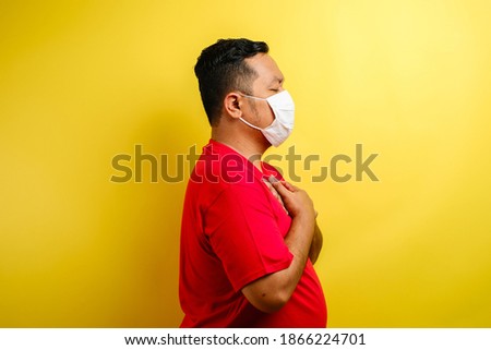 asian guy wearing mask feeling pain in his chest, gesture holding left chest. Yellow Background