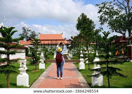 A female tourist wearing a denim shirt, a yellow hat, and a red backpack. Using a mobile phone to take pictures to capture the impression of the architecture in Phra That Sawi temple Chumphon Province