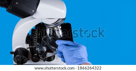 Light microscope for observation of biopsy, anatomy or histology isolated on blue background. Hand in gloves holding tissue samples. Close up of lens. Copyspace