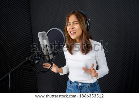 Young asian woman in white long sleeve t-shirt singing in front of black soundproofing walls. Musicians producing music in professional recording studio.
