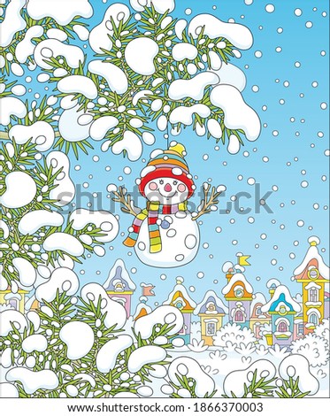 Funny toy snowman with a colorful striped scarf and a warm hat hanging on a snow-covered prickly fir branch of a Christmas tree in a snowy winter park of a pretty town