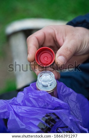 Man opens a bottle of alcoholic drink hidden in violet bag in the park. Whisky consumption outdoors. Red cap hold with two fingers. Spirits consumption in park. Close up vertical picture. Celebrate