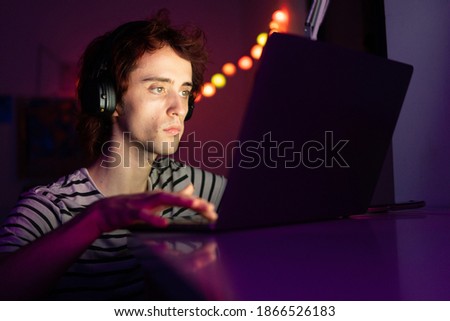 Stock photo of a young adult on his laptop indoors working. Young man on his laptop indoors playing video games wearing headphones.