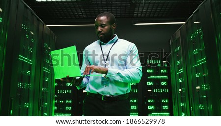 African American young man walking among servers with laptop computer and checking work of big data processors. Male analytic in data storage working on information security.