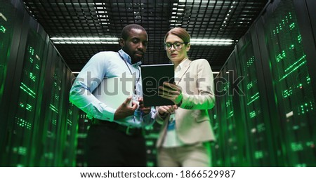 Mixed-races man and woman with tablet device discussing issue at datacenter. Multiethnic developers standing at servers storage and looking at gadget. Male and female analytics colleagues talking. Royalty-Free Stock Photo #1866529987