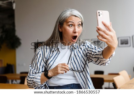 Shocked white-haired mature woman taking selfie photo on cellphone indoors