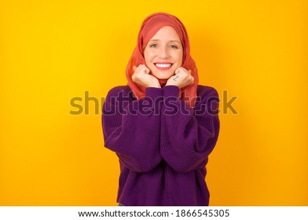 Dreamy Young beautiful muslim woman wearing hijab against yellow background keeps hands pressed together under chin, looks with happy expression, has toothy smile.