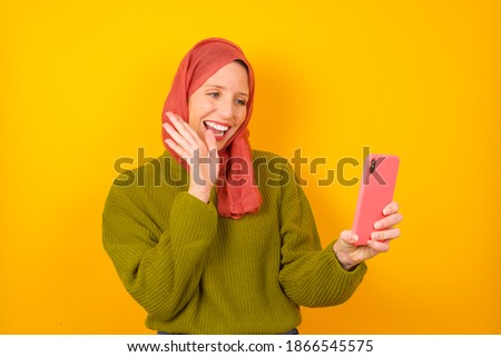 Portrait of happy friendly Young beautiful muslim woman wearing hijab against yellow background taking selfie and waving hand, communicating on video call, online chatting.