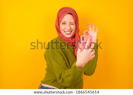 Excited Young beautiful muslim woman wearing hijab against yellow background showing smartphone blank screen, blinking eye and doing ok sign with hand.  Advertisement concept.