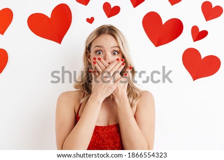 Image of a young blonde shocked surprised woman in red dress covering mouth isolated over white background with hearts