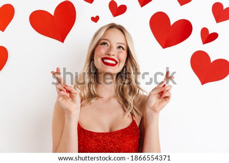 Photo of a young blonde dreaming woman in red dress with red lipstick showing crossed fingers gesture isolated over white background with hearts