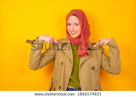 Pick me! Confident, self-assured and charismatic Young beautiful muslim woman wearing hijab against yellow background promoting oneself as wanting role smiling broadly and pointing at body.