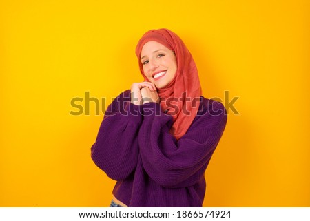 Dreamy Young beautiful muslim woman wearing hijab against yellow background with pleasant expression, closes eyes, keeps hands crossed near face, thinks about something pleasant