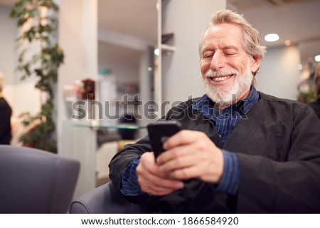 Senior Man Waiting To Have Hair Cut In Hairdressing Salon Looking At Mobile Phone