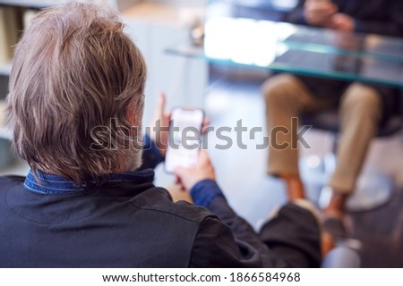 Rear View Of Senior Man Waiting To Have Hair Cut In Hairdressing Salon Looking At Mobile Phone
