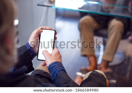 Rear View Of Senior Man Waiting To Have Hair Cut In Hairdressing Salon Looking At Mobile Phone