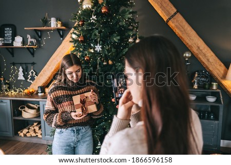 Two young women friends taking photos with gifts near Christmas tree in holidays.