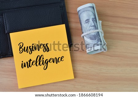 Top view of money banknote, notebook and memo note written with text BUSINESS INTELLIGENCE over wooden background. 