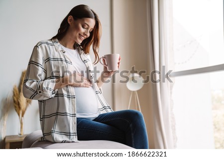 Beautiful happy pregnant woman smiling and drinking coffee while sitting on couch at home Royalty-Free Stock Photo #1866622321