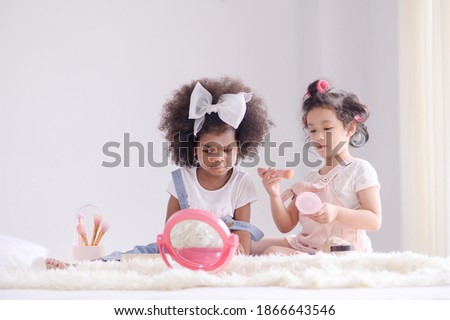 A cute little Asian girl and African friend is happily applying makeup brushes with powder in her bedroom