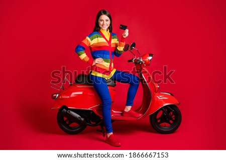 Photo portrait of confident woman holding plastic card in one hand sitting on scooter isolated on vivid red colored background