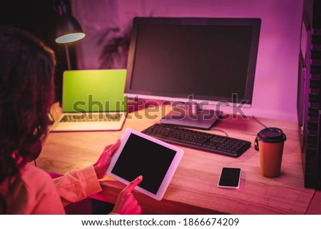 Digital tablet with blank screen in hands of gamer near computers, smartphone and coffee to go on blurred background 