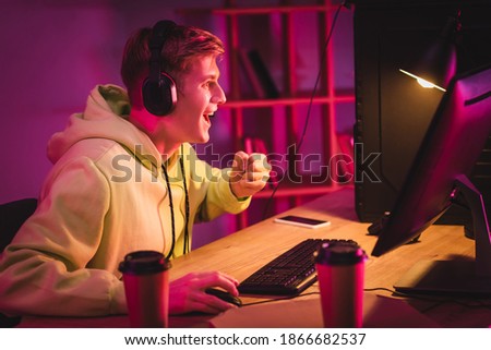 Cheerful gamer showing yeah gesture near computer, coffee to go and smartphone on blurred background 