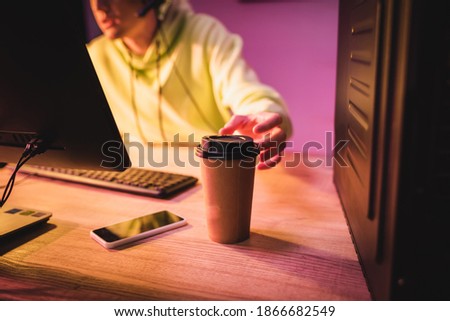 Cropped view of coffee to go and smartphone on table near gamer using computer on blurred background 