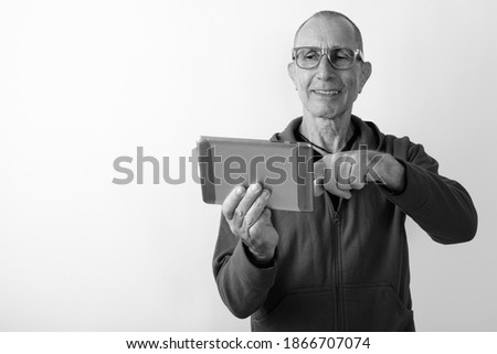 Studio shot of happy bald senior man smiling and using digital tablet while wearing eyeglasses against white background