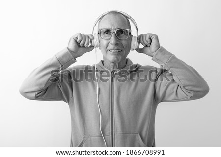 Studio shot of happy bald senior man smiling while listening to music and wearing eyeglasses against white background