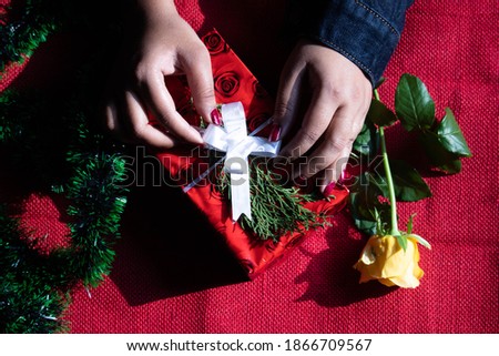 Hands Of Female Girl In Blue Jeans Jacket Packing Gift Box Wrapped In Red Paper Silver Ribbons With Green Christmas Fur Tree Leaves Tinsel Yellow Rose Around On Maroon Burlap Jute Fabric