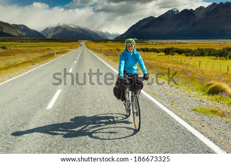 A mountain biker rides along a winding asphalt track in a valley among fields on a background of blue sky with clouds in New Zealand 