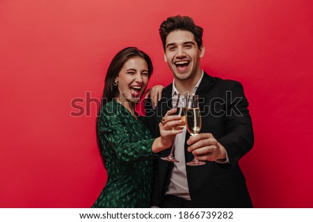 Cheerful young pair of people in holiday outfits, smiling, holding glasses with champagne and looking into camera isolated on red background 
