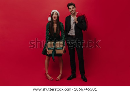 Pretty young girl in dark dress and Santa Claus hat holding gift box and standing with handsome young man, wearing black suit, light shirt and smiling. People posing against red plain background 