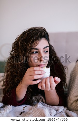 portrait of a white woman with curly brunette hair lying on the bed with a glass of coffee