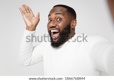 Close up of surprised excited young african american man in casual basic sweater doing selfie shot on mobile phone spreading hands looking camera isolated on white color background studio portrait