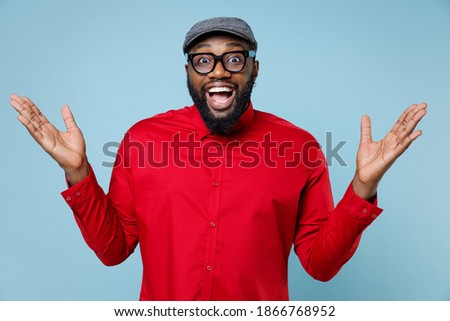 Surprised young bearded african american man 20s wearing casual red shirt cap eyeglasses standing keeping mouth open spreading hands isolated on pastel blue color wall background studio portrait