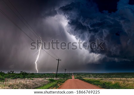 A nighttime, tornadic mezocyclone lightning storm shoots bolt of electricity to the ground and lights up the field and dirt road in Tornado Alley.

 Royalty-Free Stock Photo #1866769225
