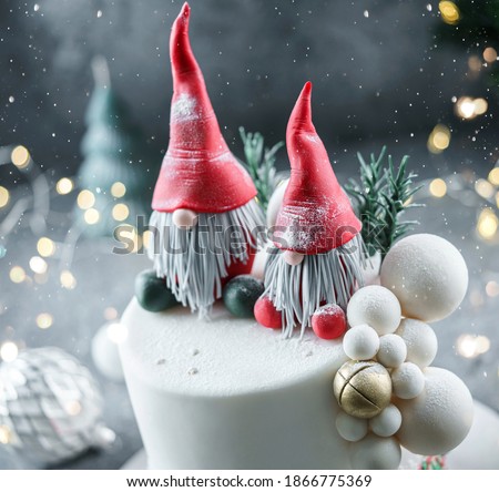 Christmas cake decorated with sweet figures Santa's gnomes, fir branches and bells on bright background with garland, candy, bokeh. Winter baking at Xmas or New Year holiday, close up