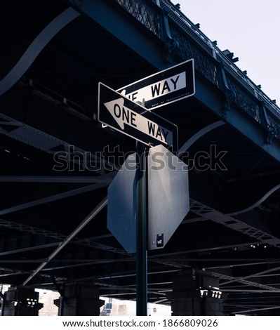 Two 'One Way' signs intersecting under a bridge in dramatic light in New York City