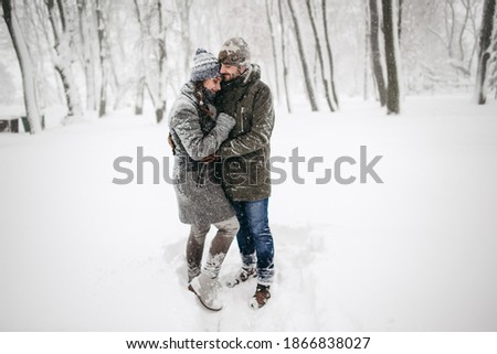 A young beautiful European couple in a gray coat, knitted hat and mittens walks in a winter park during a snowfall, having fun, hugging, running. Snow, nature, trees, man and woman, girl celebrating.