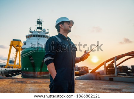 Marine Deck Officer or Chief mate on deck of offshore vessel or ship , wearing PPE personal protective equipment - helmet, coverall. He holds VHF walkie-talkie radio in hands. Royalty-Free Stock Photo #1866882121