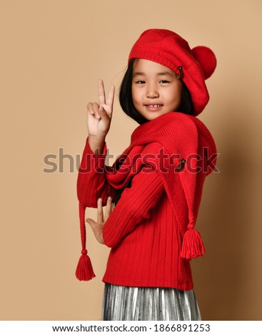Little asian girl in red hat, scarf and sweater shows victoria victory sign with her fingers. Place for text or advertising.