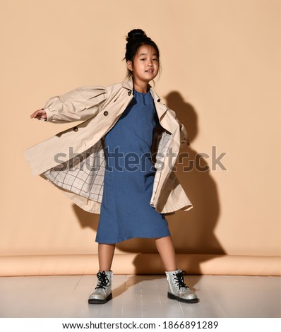 small Korean brunette wearing a blue dress, beige trench coat and boots is spinning. Children's fashion concept.