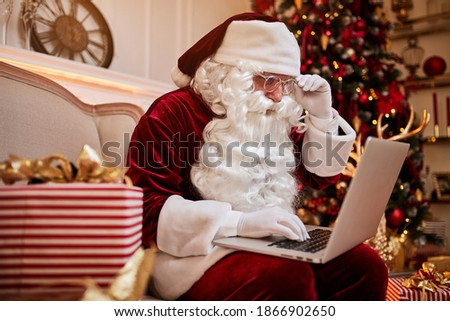 Santa Claus sitting at his home and reading email on laptop with сhristmas requesting or wish list near the fireplace and tree with gifts. New year and Merry Christmas , happy holidays concept