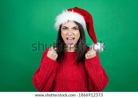 Young beautiful woman wearing Christmas Santa hat over isolated green background very happy and excited making winner gesture with raised arms, smiling and screaming for success.