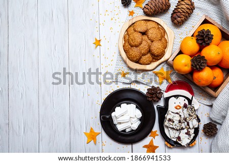Cozy winter still life with tangerines, cookies, chocolate, pine cones and a cup of marshmallows on a light wooden table. Top view, place for text.