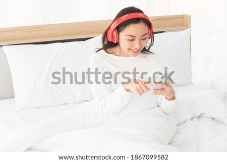 Happy smile, Relaxing Joyful young beautiful Asian woman using smartphone and listening to music with red wireless headphones and sitting on a white bed in bedroom at home. Chill out, leisure concept.