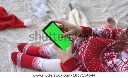 Brunette woman is sitting on a fluffy blanket uses mobile phone with Green Screen for Copy Space between Christmas decorations. Christmas and New Year Celebration Concept.