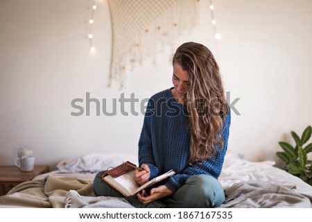 Woman in blue sweater sat on her bed writing in her leatherbound journal Royalty-Free Stock Photo #1867167535